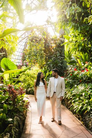 A couple walks hand in hand through a lush greenhouse pathway filled with tropical plants and bright natural light.