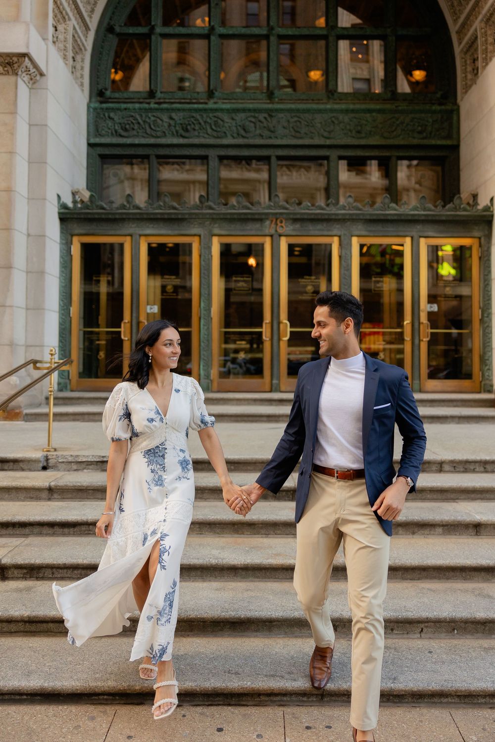 A couple sits together on stone steps in front of a historic building with large windows.