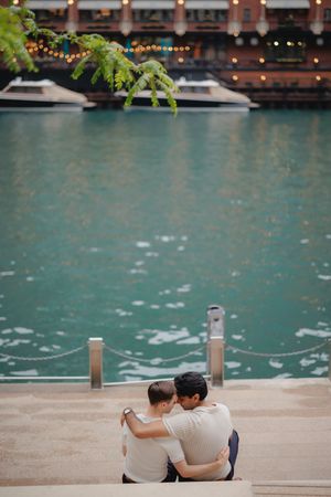 A couple sits together on steps by turquoise waters with string lights and greenery overhead at a waterfront location.