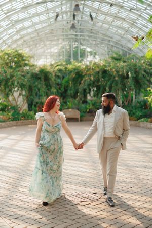 A couple walks hand in hand through a sunlit greenhouse conservatory with a glass ceiling and lush greenery.