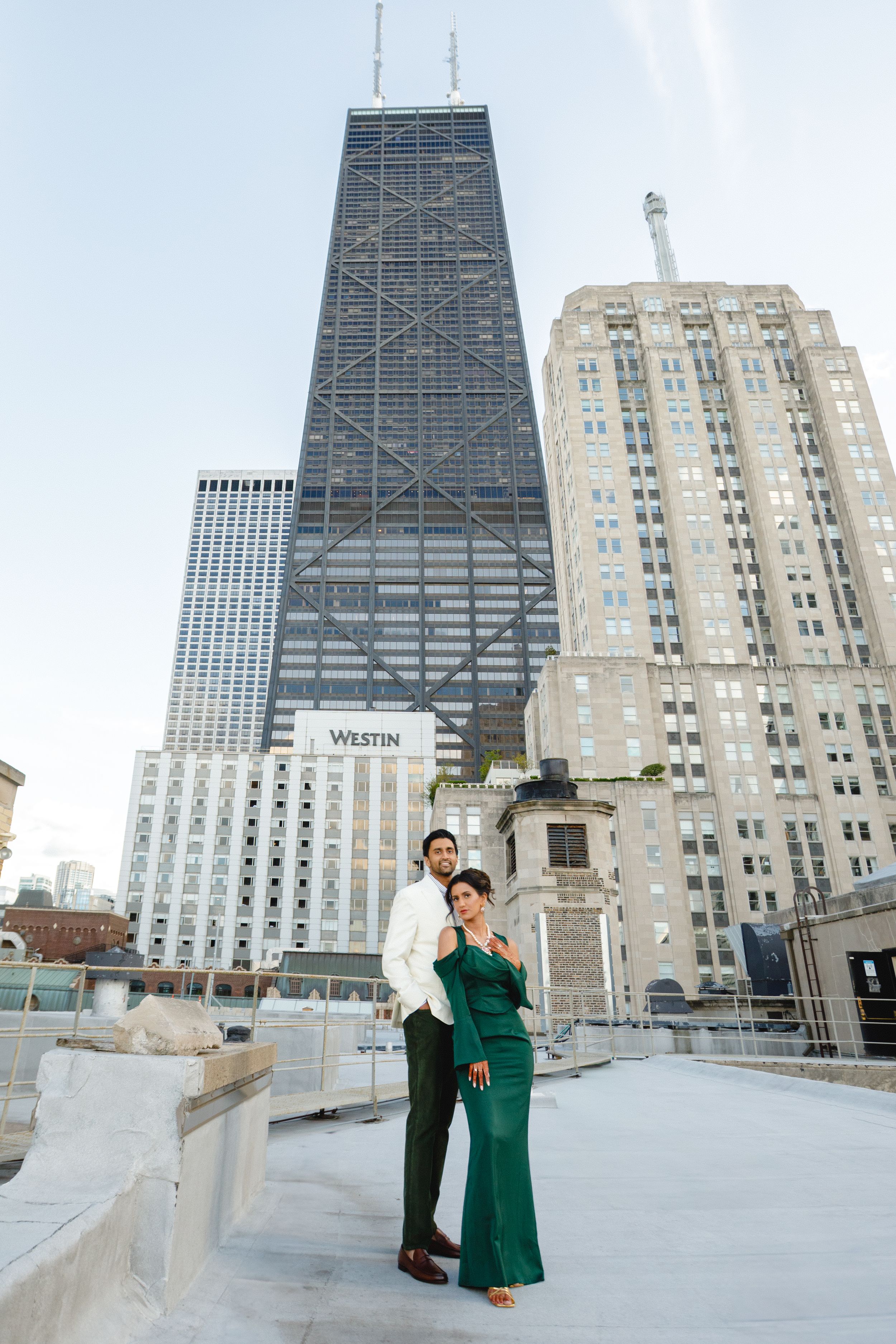 A couple stands on a rooftop terrace with Chicago's iconic John Hancock building and city skyline in the background.