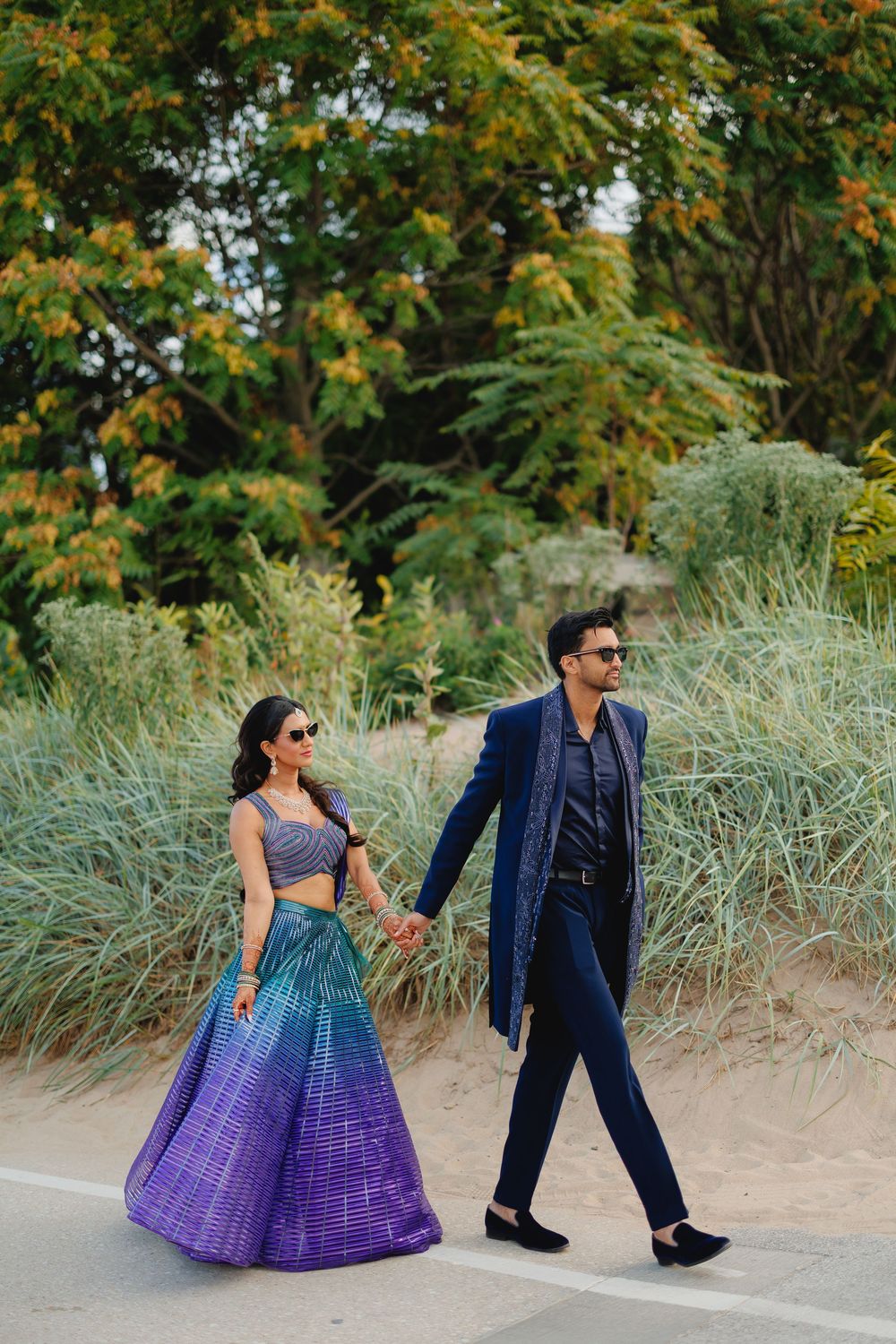 A couple walking outdoors in formal attire with a blue and purple gradient lehenga and navy suit against natural foliage.