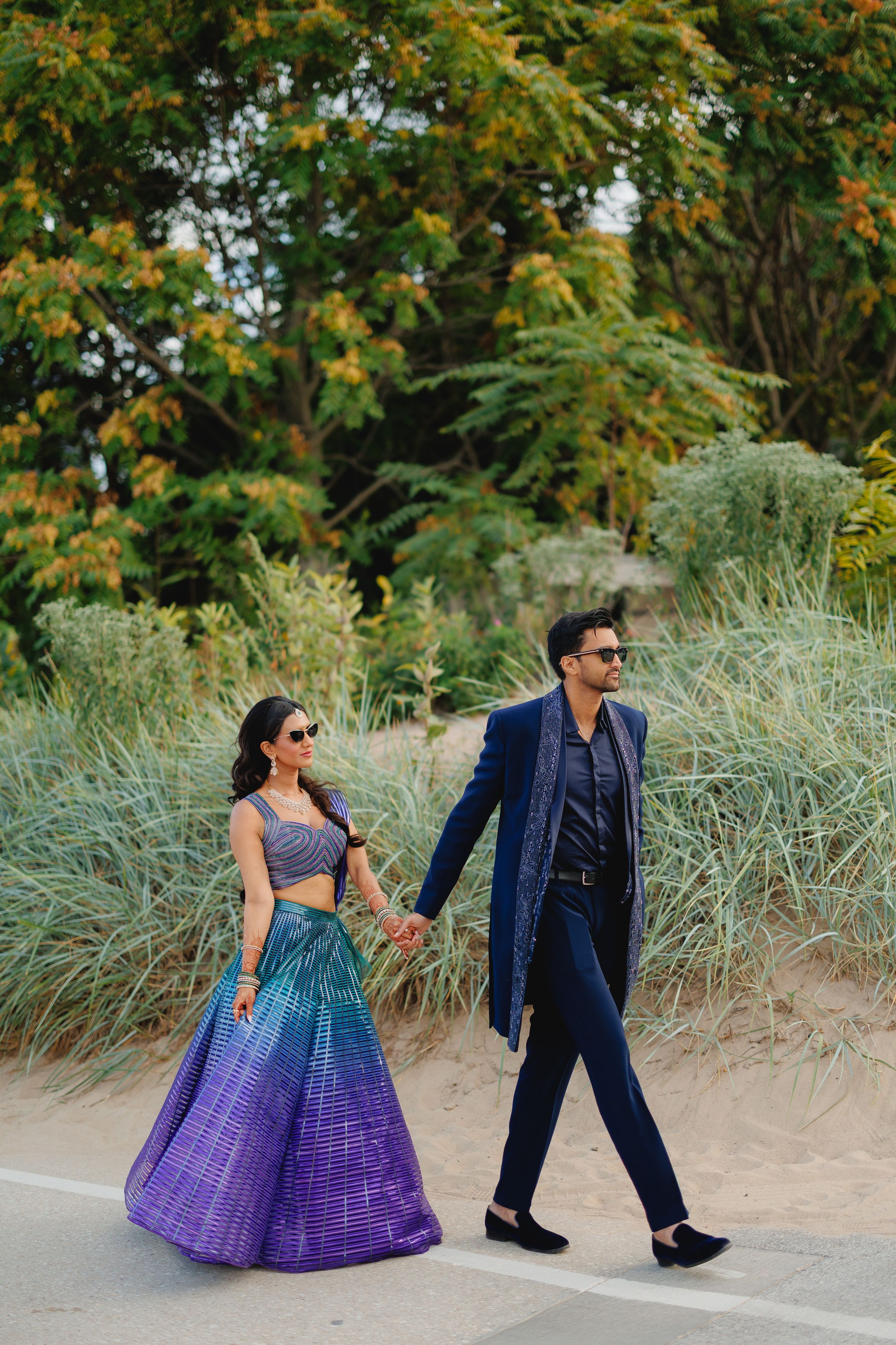 A couple walking outdoors in formal attire with a blue and purple gradient lehenga and navy suit against natural foliage.