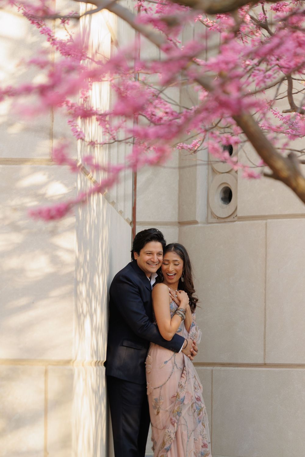 Couple embraces under blooming cherry blossom branches against white wall in romantic spring moment.
