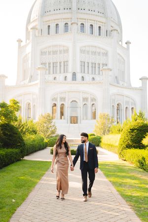 A couple walks hand in hand on a path leading to the majestic white domed Bahai Temple on a sunny day.