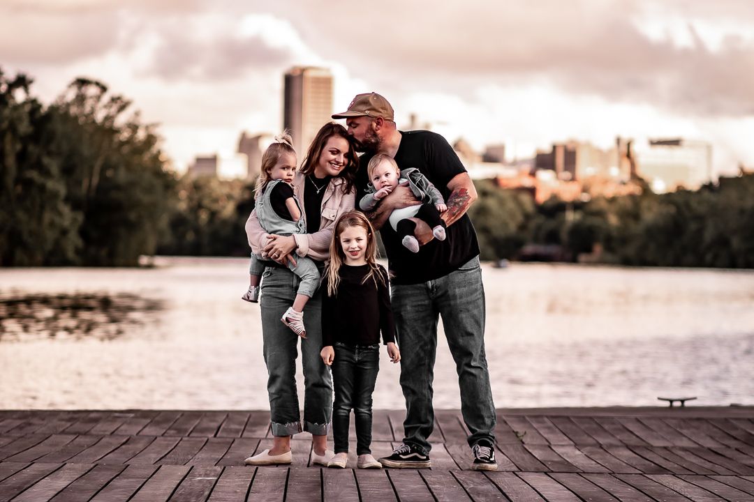 Happy family of five posing on a wooden dock with a city skyline and lake in the background.