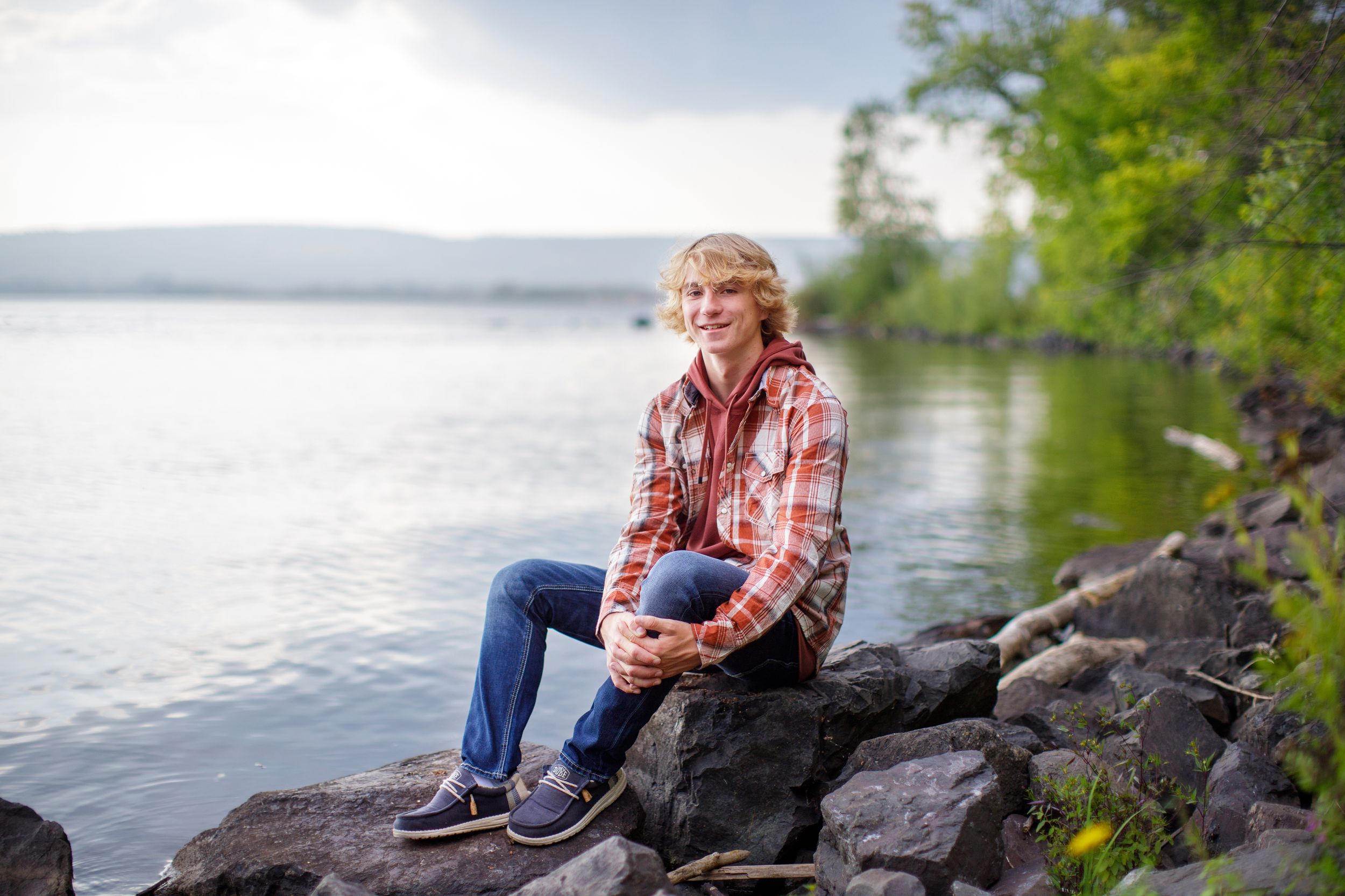 Person in plaid shirt and jeans sits peacefully on rocky shoreline overlooking serene lake on cloudy day.