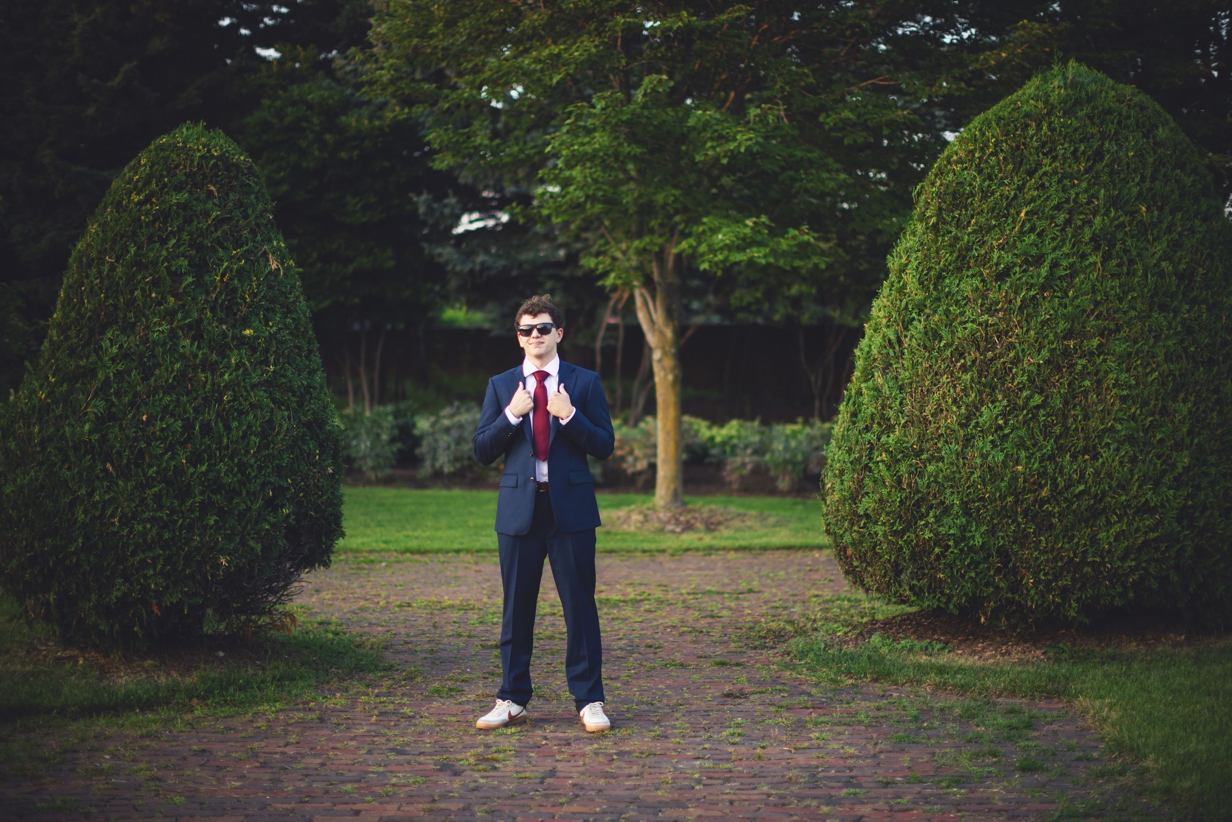 A person in a dark suit stands in a park-like setting surrounded by green trees and manicured hedges.
