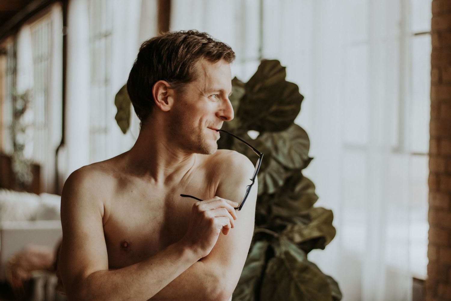 Shirtless smiling man holding glasses near a bright window with tropical plants in a cozy indoor setting.