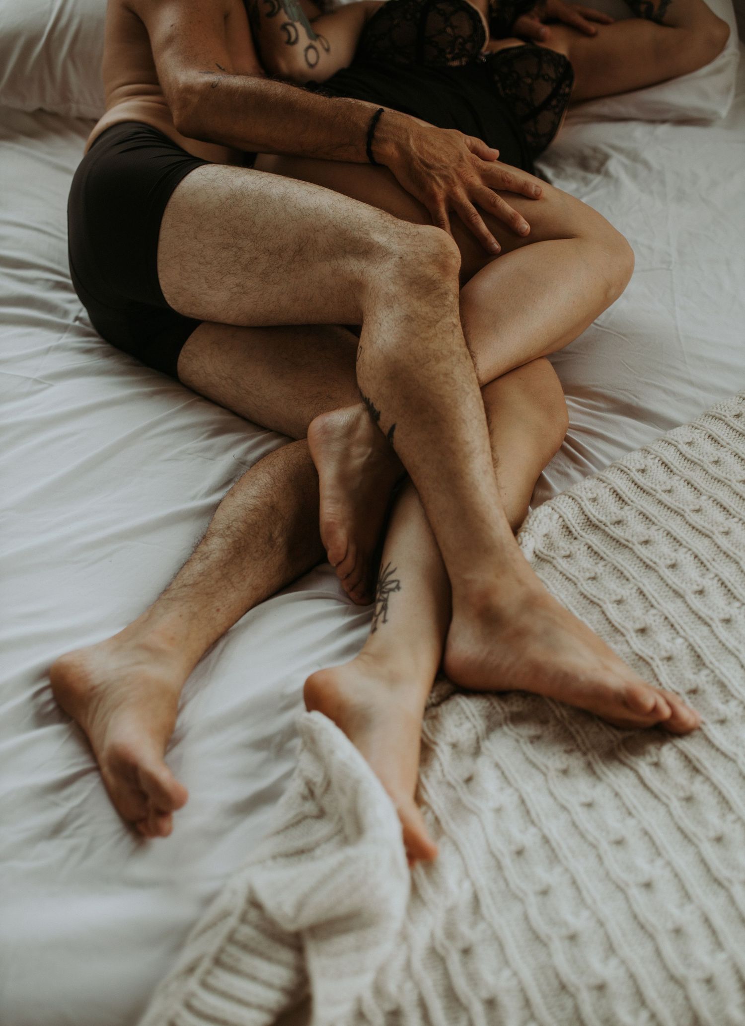 Couple relaxing together on white bedding, intertwined legs visible, cozy intimate bedroom setting.