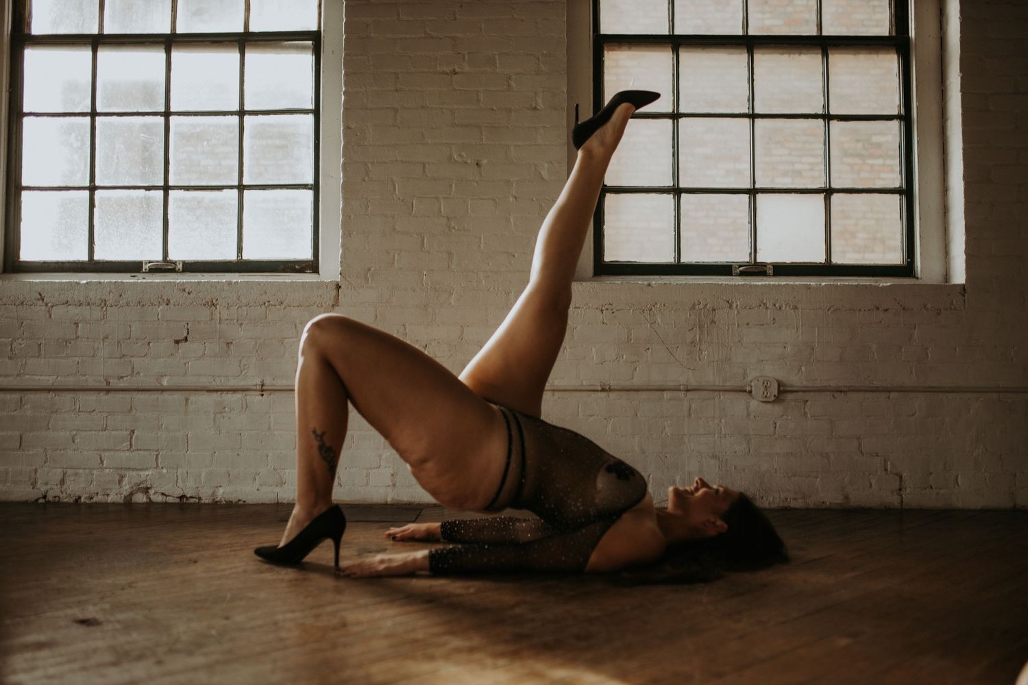 Artistic photo of person in black lingerie doing a bridge pose in industrial space with windows.