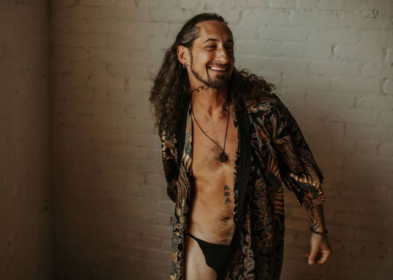 Smiling man with long curly hair wearing an open patterned robe and necklace, posing against a white brick wall.