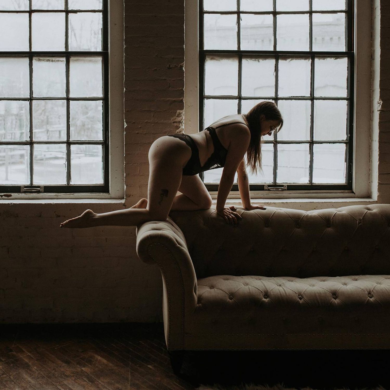 Woman in lingerie posing on a tufted sofa near large industrial windows in a moody loft setting.