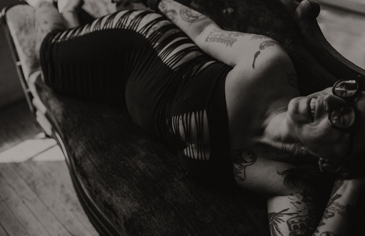 Black and white photo of tattooed woman reclining on vintage sofa in black dress.
