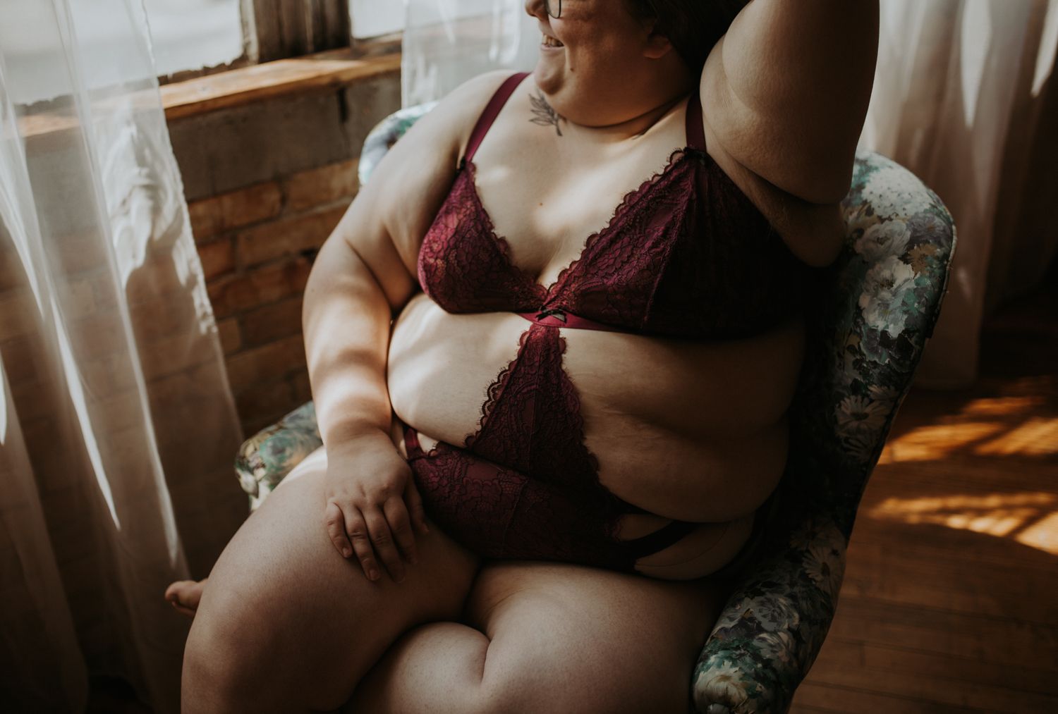 Plus-size woman in burgundy lace lingerie set posing confidently in a floral armchair by a sunlit window.