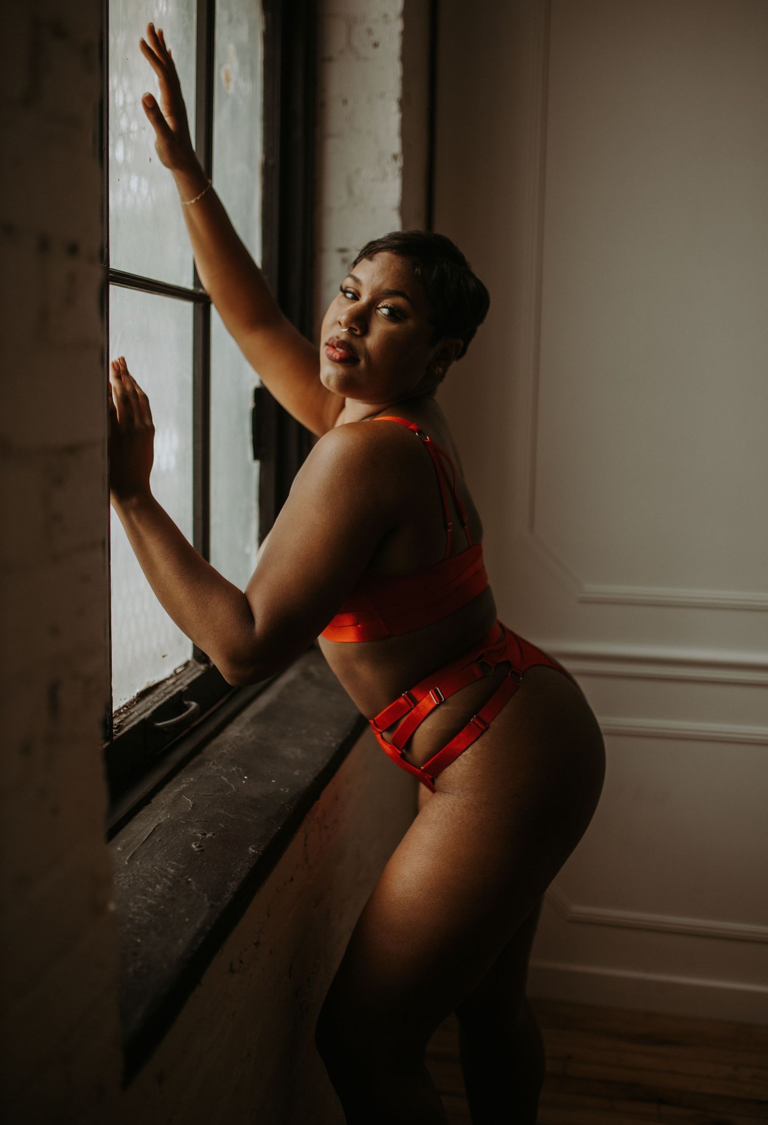 Woman in red lingerie posing by a window in a dimly lit room, looking over her shoulder.