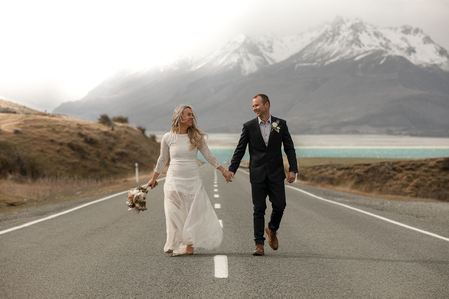 A couple walks down an empty road with snow-capped mountains and turquoise lake in the background.