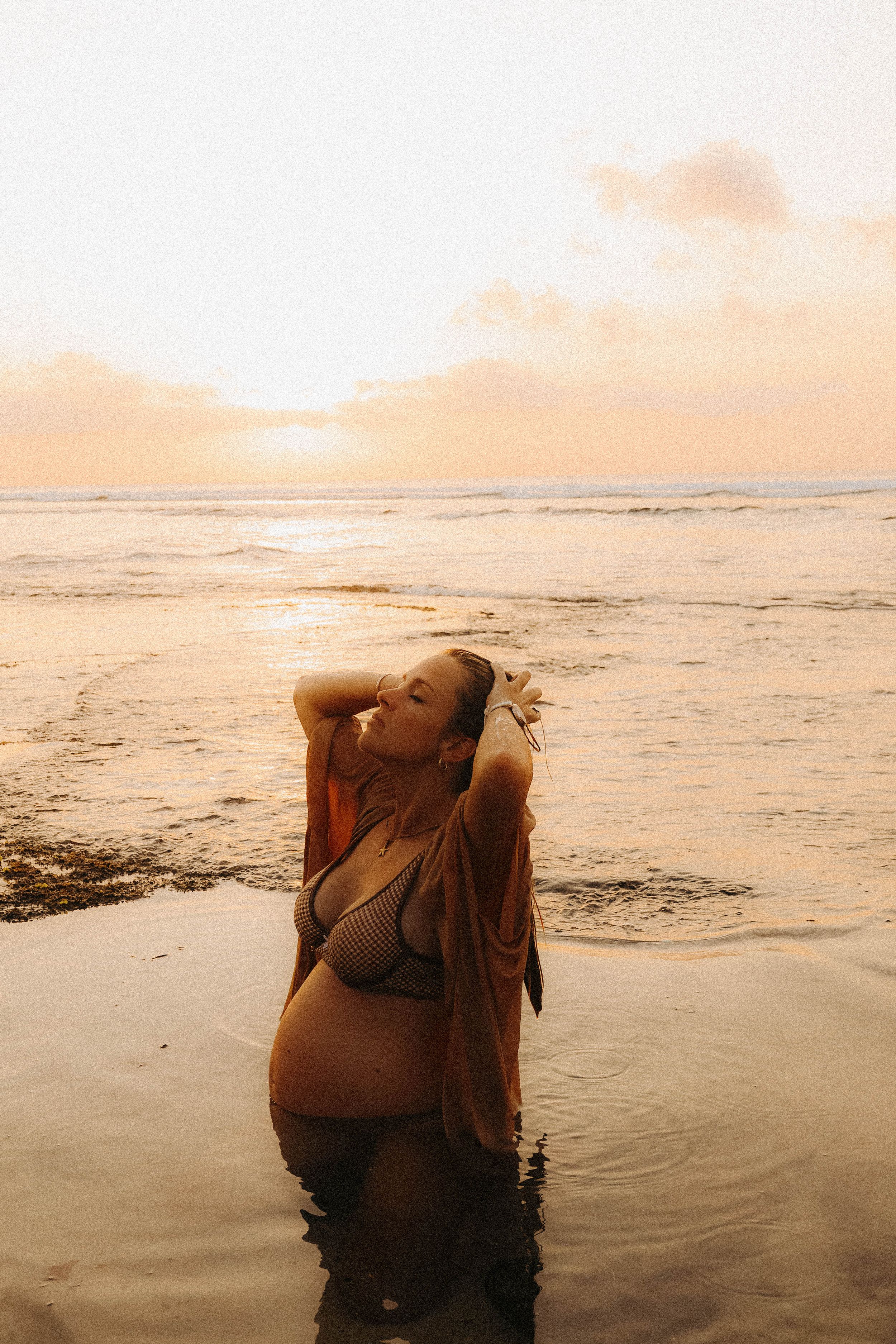Pregnant woman in bikini standing in ocean water during stunning sunset.