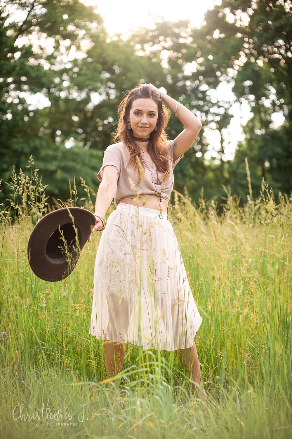 Woman in white floral skirt holding cowboy hat poses in sunlit meadow with tall grass and trees.