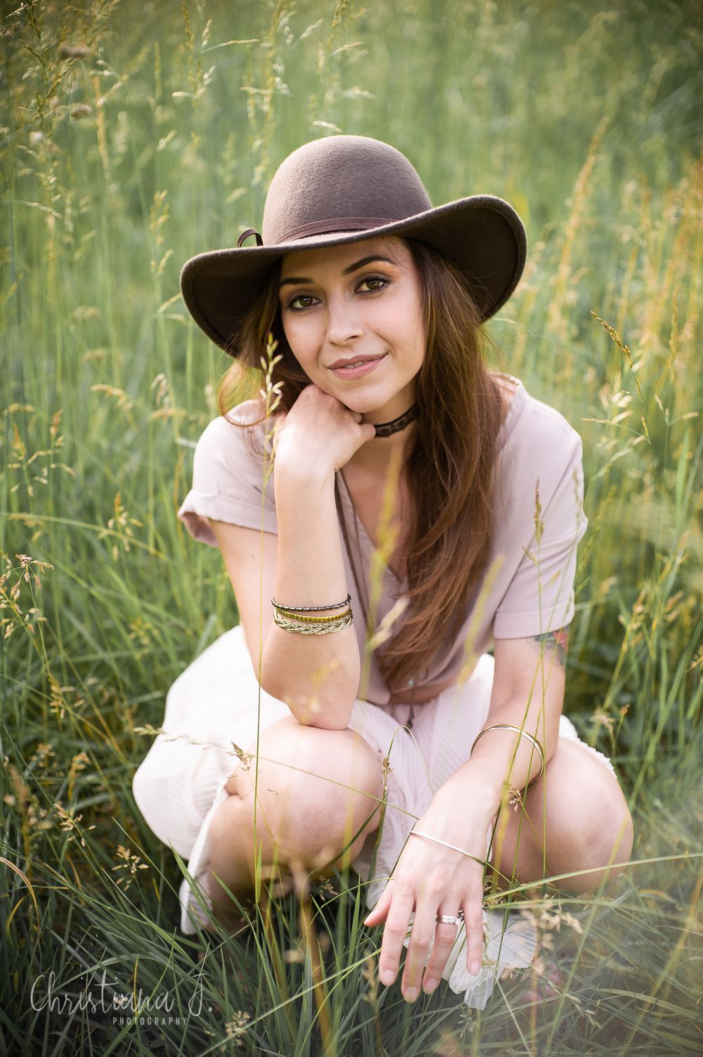 Young woman in wide-brim hat and white dress crouching in tall green grass, smiling softly for a portrait photo.