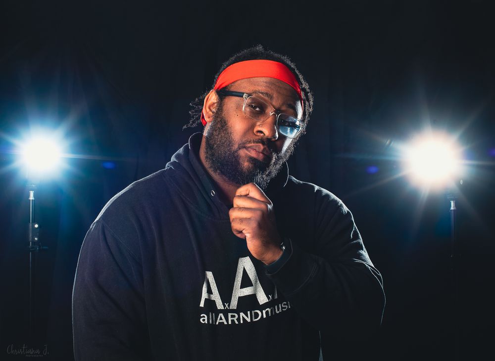 A man in a red headband and black hoodie poses confidently under stage lights in a professional music photo shoot.