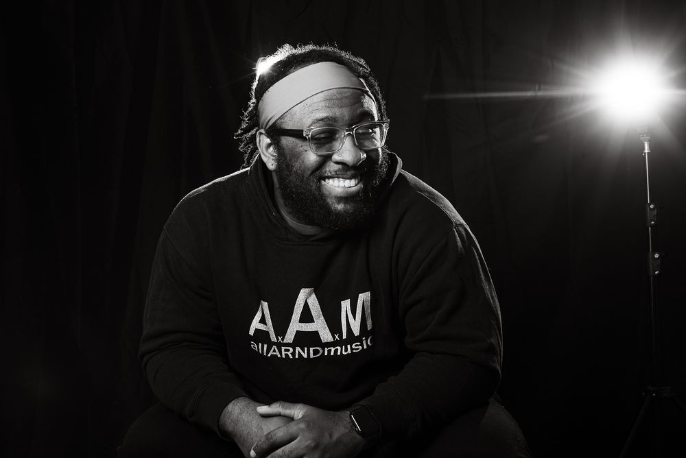 Smiling bearded man wearing glasses and AAM sweatshirt, photographed in black and white under studio lighting.