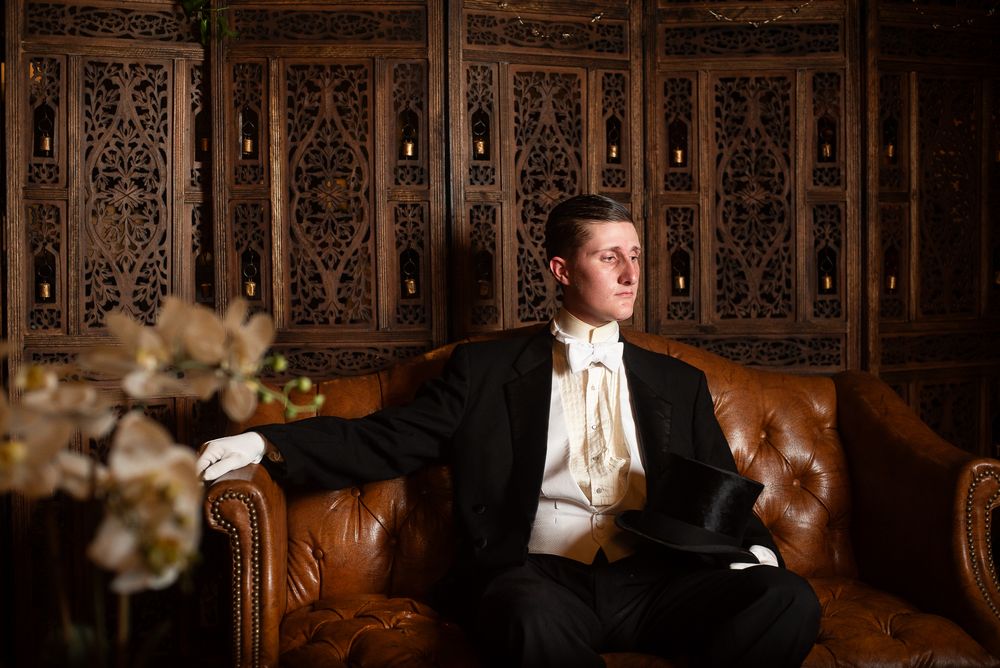 Elegant man in formal black tuxedo and white cravat sitting on leather sofa holding top hat in ornate room.
