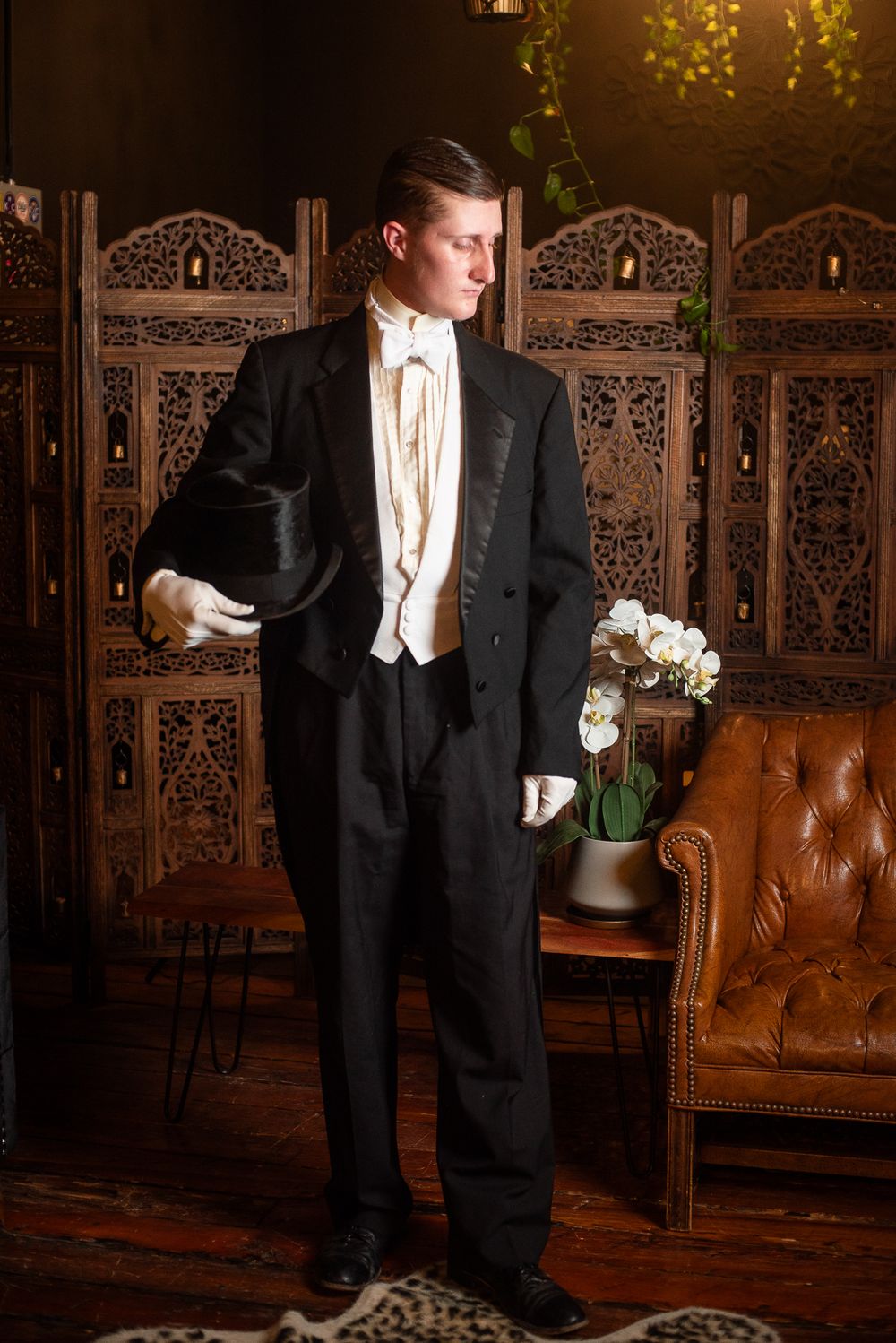 Man in formal black tailcoat suit with white gloves and top hat poses in elegant vintage-styled room with leather chair.