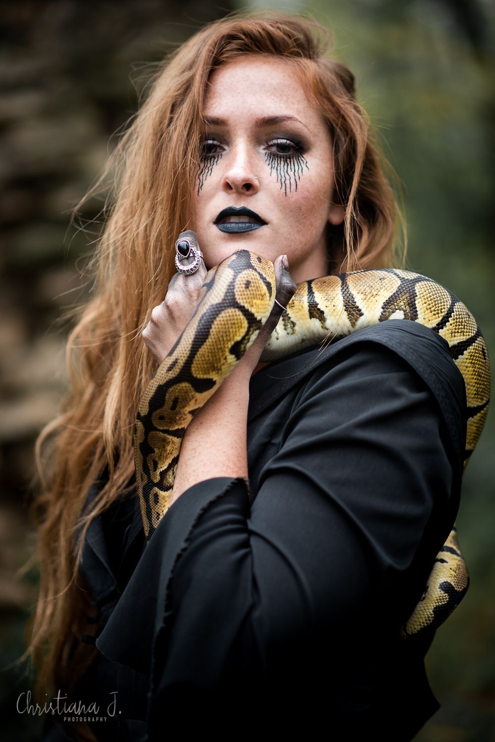 Woman with dramatic black lipstick and crystal eye makeup holds a ball python snake, wearing a black jacket outdoors.
