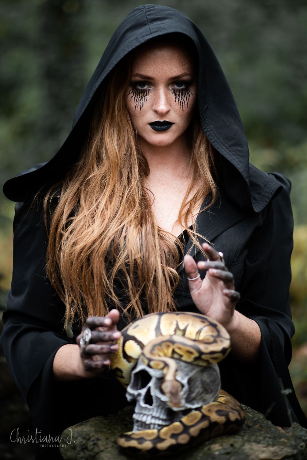 Woman in witch costume with black makeup holds hands over a skull with a snake in a dark forest setting.