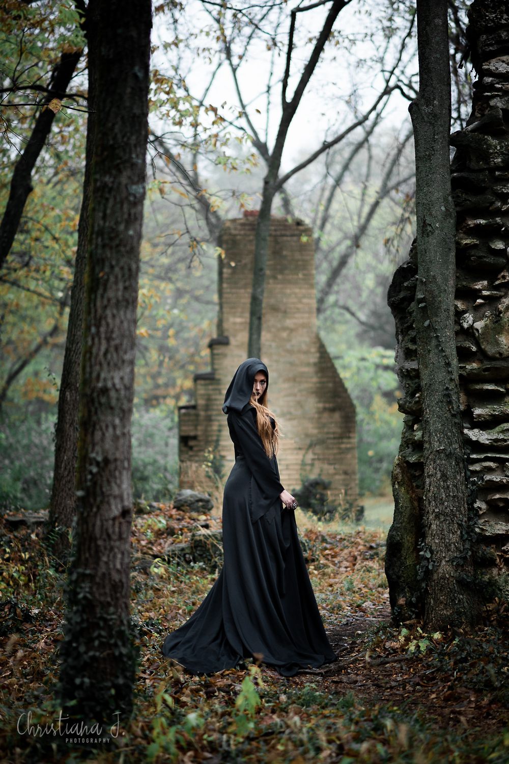Woman in black hooded gown stands among autumn trees near stone ruins in a dark, mysterious forest setting.