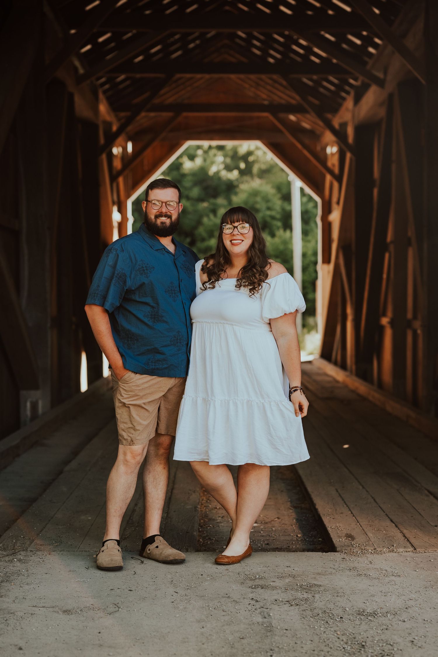 Couple posing inside a rustic wooden covered bridge, man in blue shirt and woman in white dress smiling.