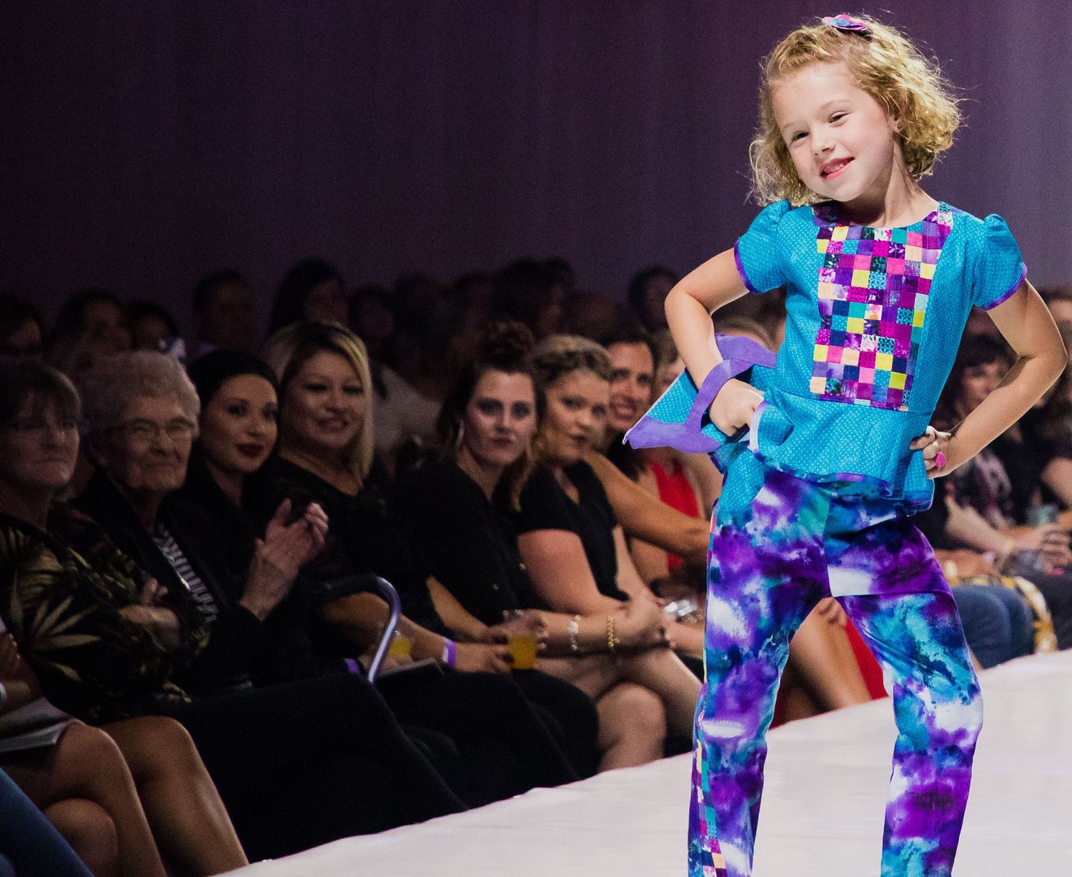 A young model strikes a pose on the runway wearing a colorful tie-dye outfit with turquoise and purple patterns.
