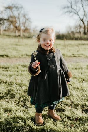 A small child wearing a dark winter coat plays joyfully in a sunlit grassy field on a bright autumn day.