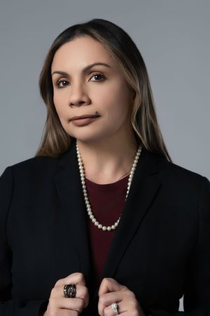 Professional business headshot photo of a woman holding her black blazer jacket over a burgundy top  in front of a gray background