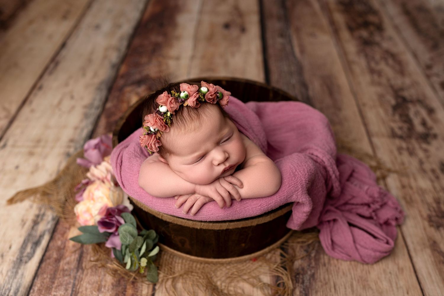 Newborn peacefully resting in pink wrap on wooden bowl with floral crown and rustic wooden floor background.