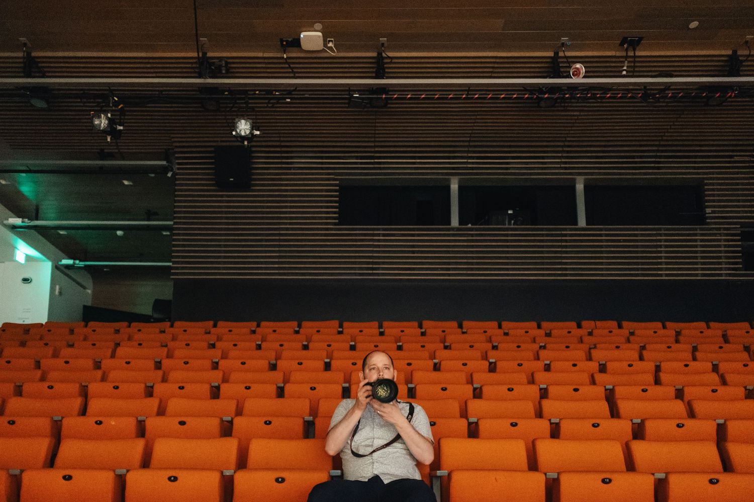 Lone man sitting in empty orange theater seats holding a camera in a modern auditorium.