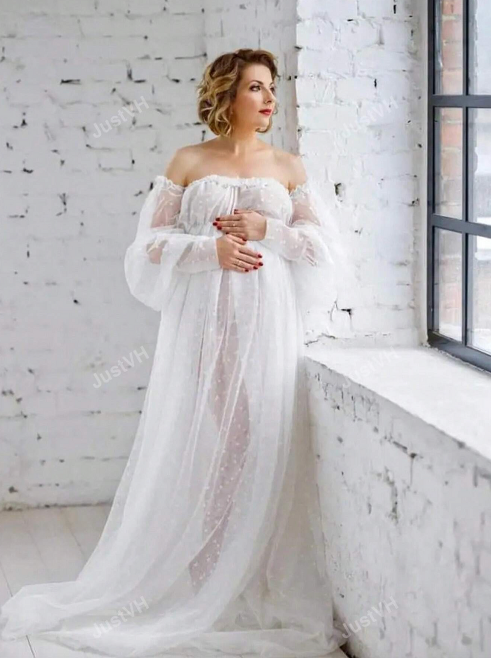 A bride in an ethereal off-shoulder white gown with billowing sleeves poses by a window in a rustic white brick room.