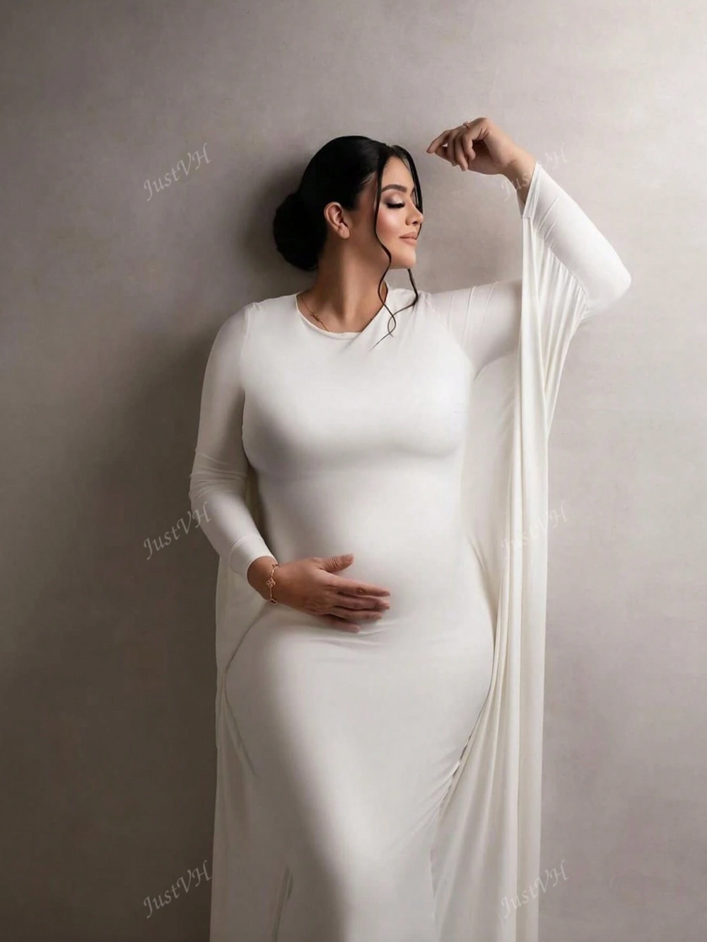A graceful pregnant woman in a flowing white dress poses against a neutral wall in an elegant maternity portrait.