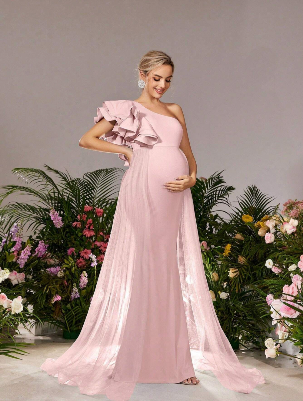 A flowing pink one-shoulder maternity gown with ruffles surrounded by tropical flowers and greenery.