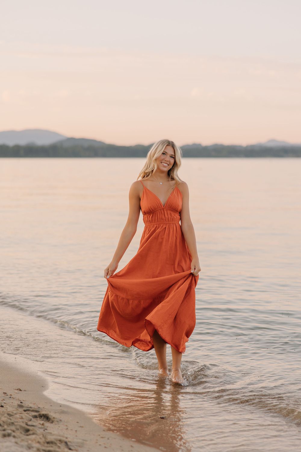 Woman in flowing orange dress walking along the water's edge at sunset.