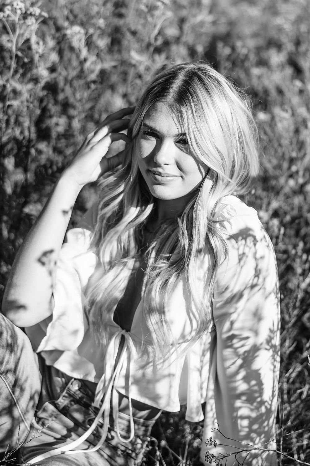 Black and white portrait of a person smiling outdoors with long blonde hair and a white top.