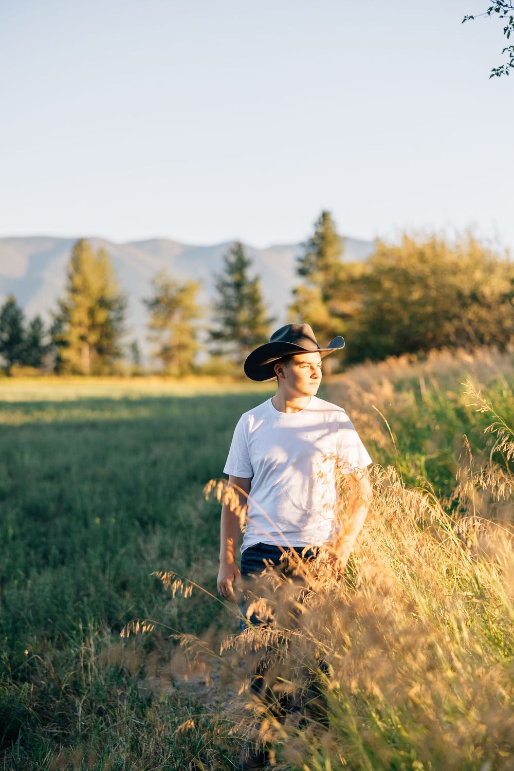 high school seniorvin a white t-shirt and hat standing in a golden field with mountains in the background.