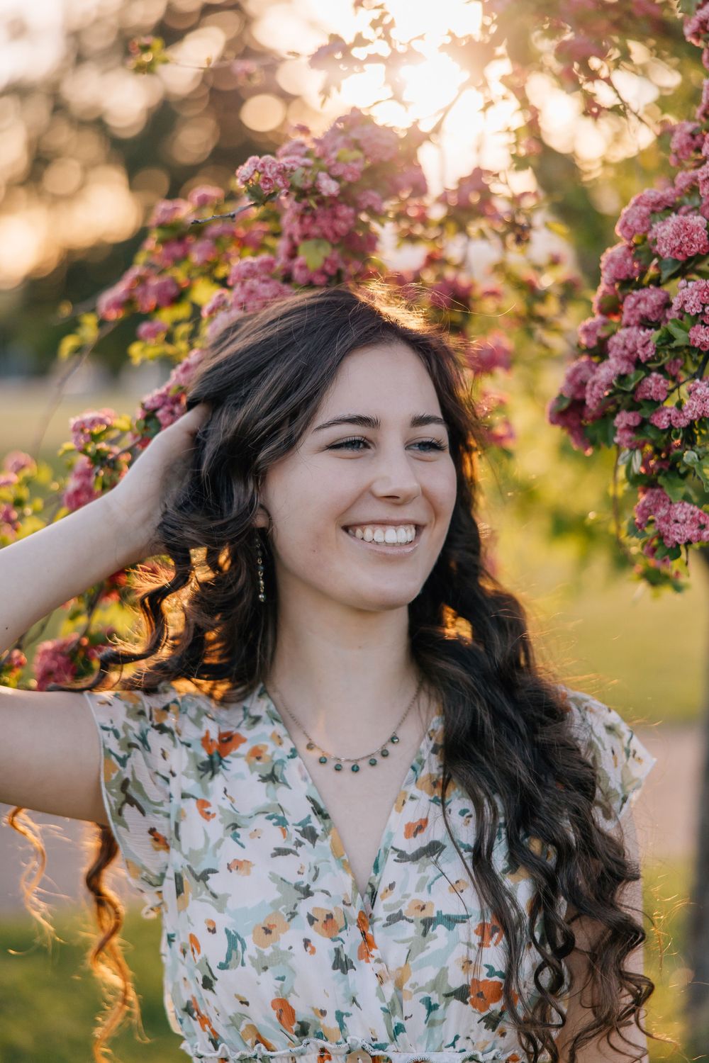 Portrait of a high school senior with long dark hair smiling in front of pink flowers at sunset.