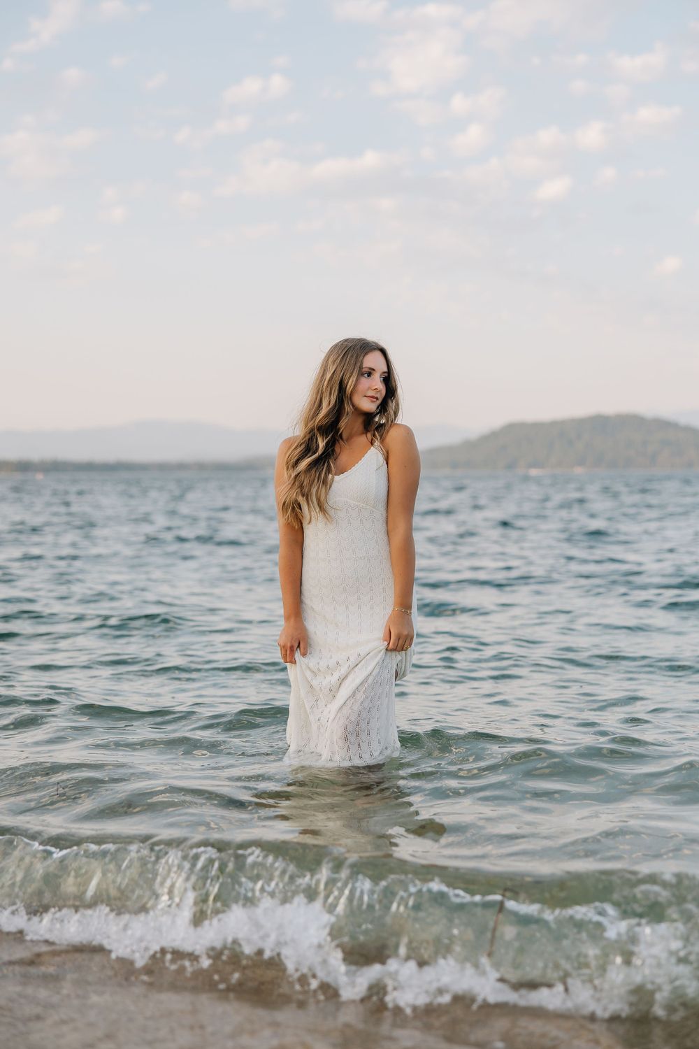 Woman in white lace dress standing in ocean waves at sunset.