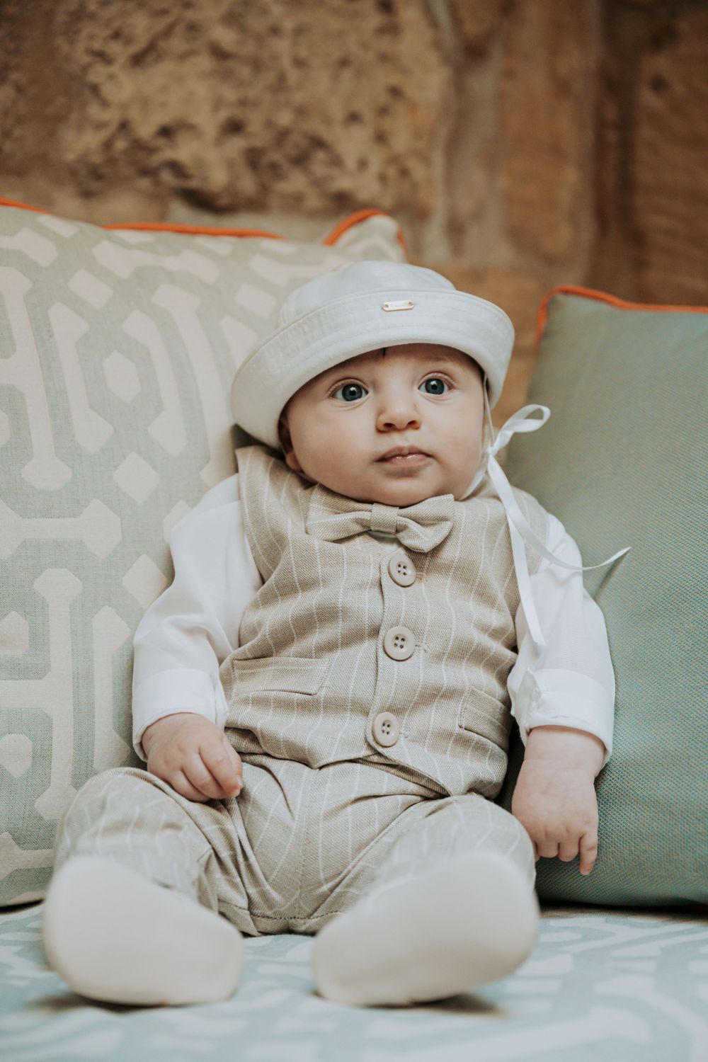 Infant in beige outfit and white cap resting on mint green cushion.