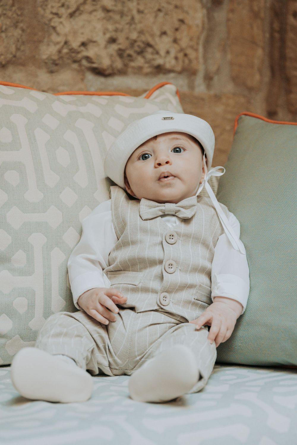 Baby wearing white outfit and cap lying on soft cushions.