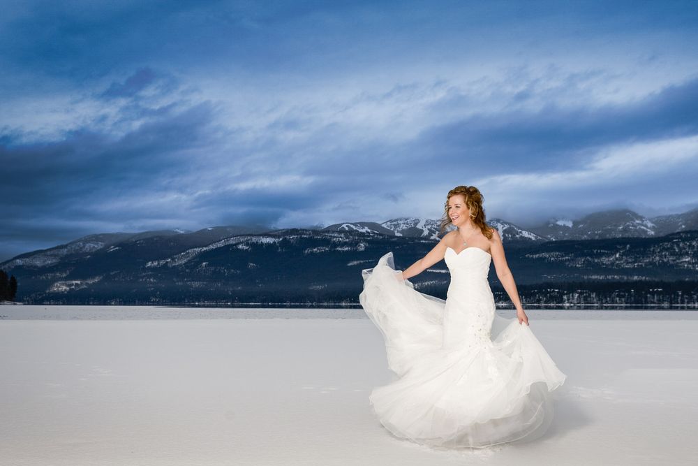 A bride in a flowing white dress poses against a snowy mountain landscape with dramatic blue skies.