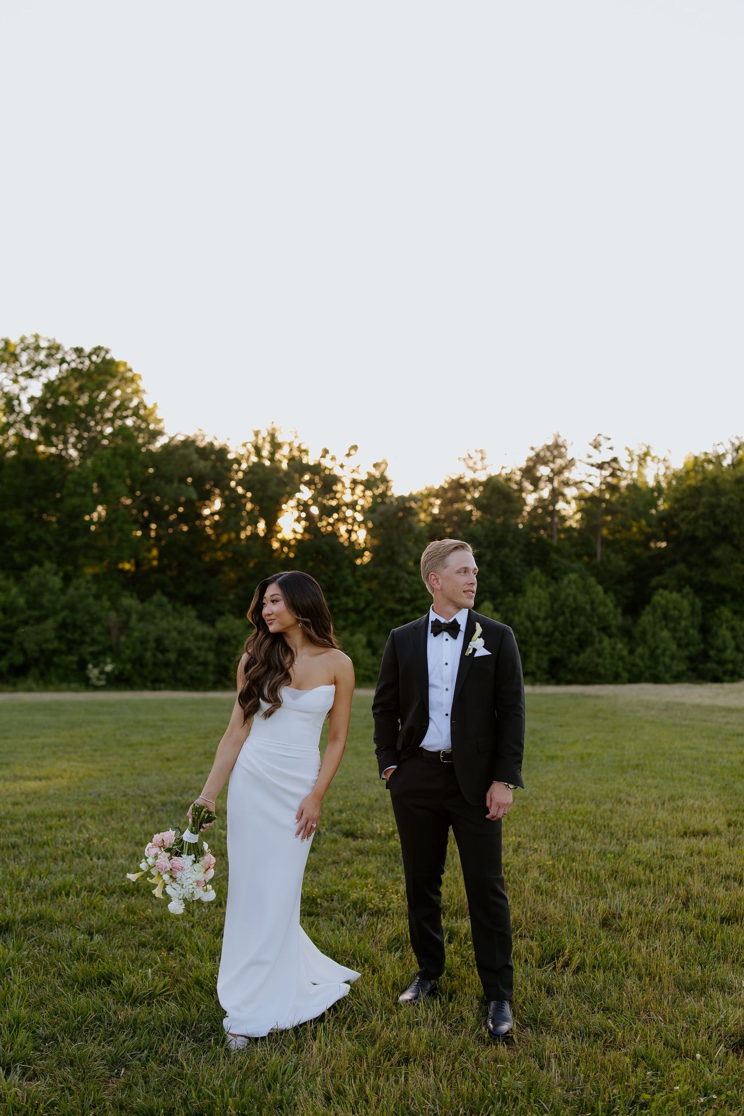 A couple walks through a grassy field at sunset in formal wedding attire.