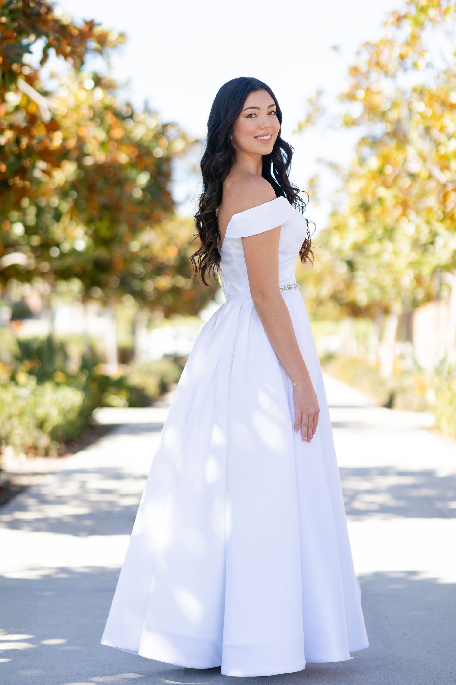 A high school senior in a flowy white dress smiles while standing on a walkway near a Mediterranean-style building with cypress trees.